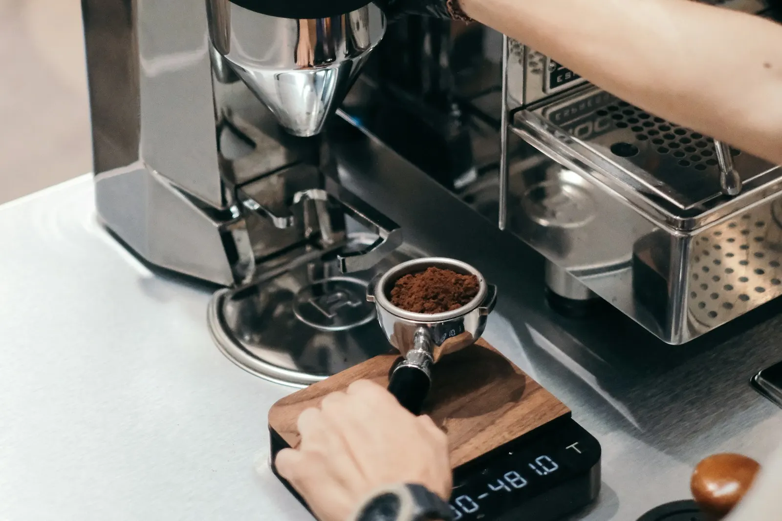 A hand resting a portafilter of fresh grounds on a wooden-topped digital scale beside a grinder